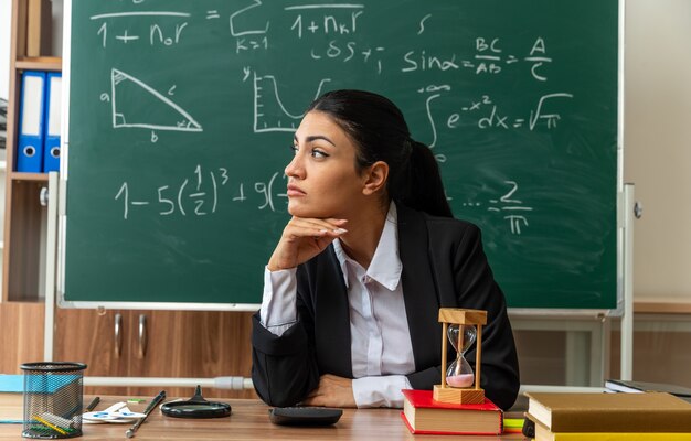 looking side young female teacher sits at table with school supplies putting hand under chin in classroom