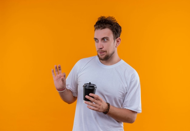 Looking at side young guy wearing white t-shirt holding cup of coffee showing okay gesture on isolated orange background