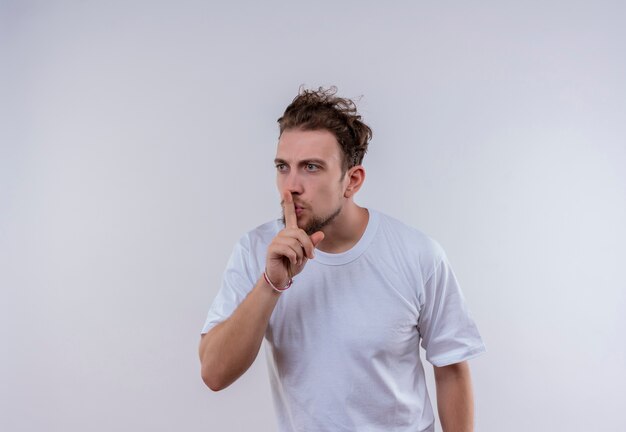 Looking at side young guy wearing white t-shirt showing silence gesture on isolated white background