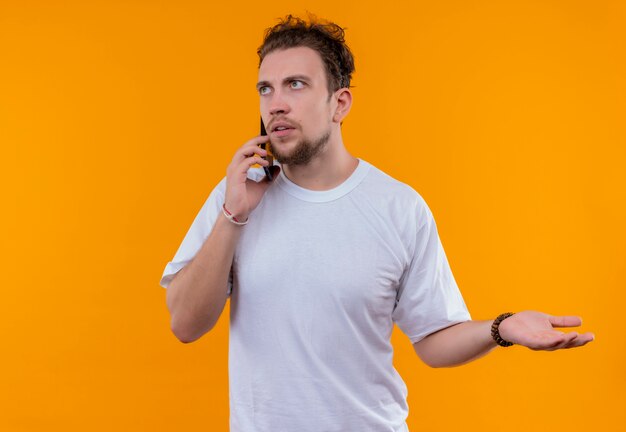 Looking at side young guy wearing white t-shirt speaks on phone holding out hand to side on isolated orange background