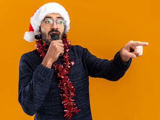 Looking at side young handsome guy wearing christmas hat with garland on neck speaks on microphone points at side isolated on orange background with copy space