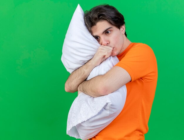 Looking at side young ill man hugged pillow putting hand on mouth isolated on green background