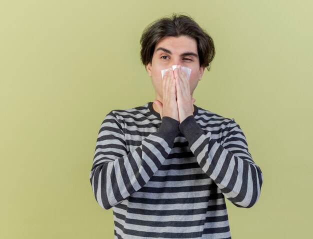 Looking at side young ill man wiping nose with napkin isolated on olive green background
