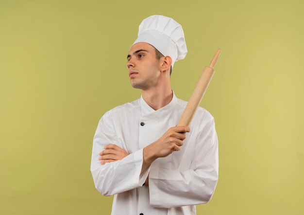 Looking at side young male cook wearing chef uniform crossing hands holding rolling pin on isolated green wall with copy space