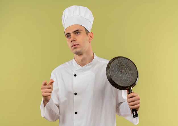 Глядя на сторону молодого повара-мужчину в униформе шеф-повара, держащего сковороду и показывающего вам жест на изолированной зеленой стене