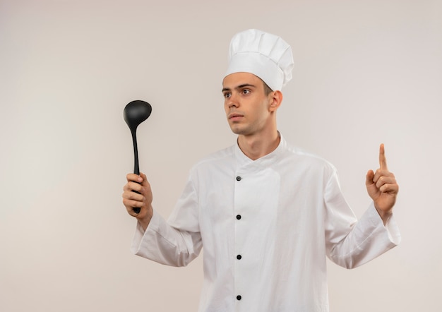 Looking at side young male cook wearing chef uniform holding ladle points finger to up on isolated white wall