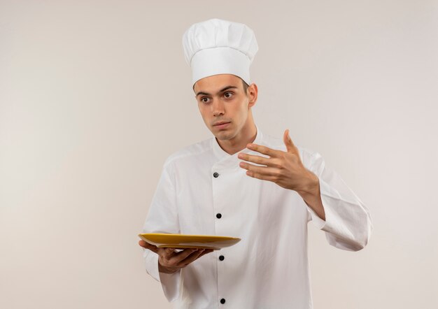 孤立した白い壁に彼の手で皿に何かを持っているふりをしているシェフの制服を着た若い男性料理人の側を見て