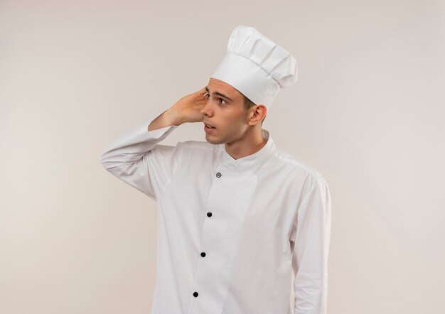 Looking at side young male cook wearing chef uniform putting hand on ear on isolated white wall