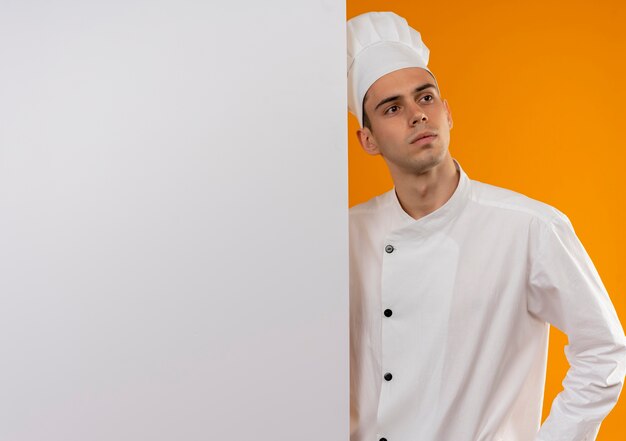 Looking at side young male cool wearing chef uniform holding white wall on isolated yellow wall with copy space