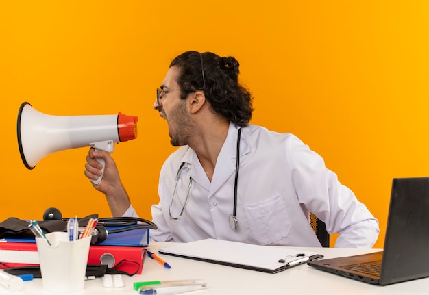 Foto gratuita guardando al lato giovane medico maschio con occhiali medici che indossano accappatoio medico con stetoscopio seduto alla scrivania