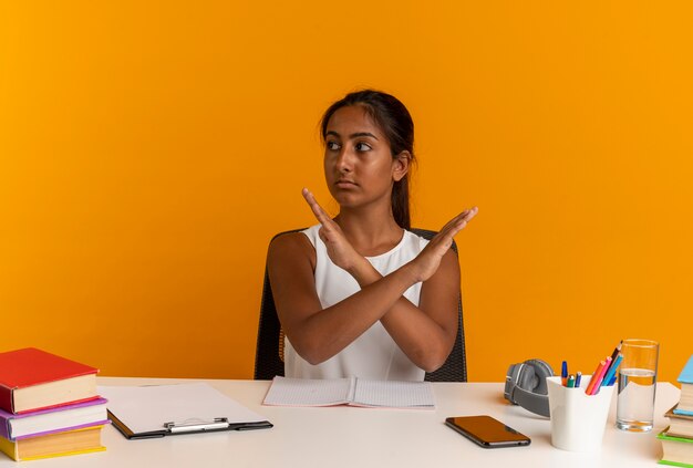 Looking at side young schoolgirl sitting at desk with school tools showing gesture of no