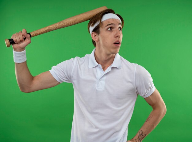 Free photo looking at side young sporty guy wearing headband with wristband putting baseball bat on head isolated on green wall