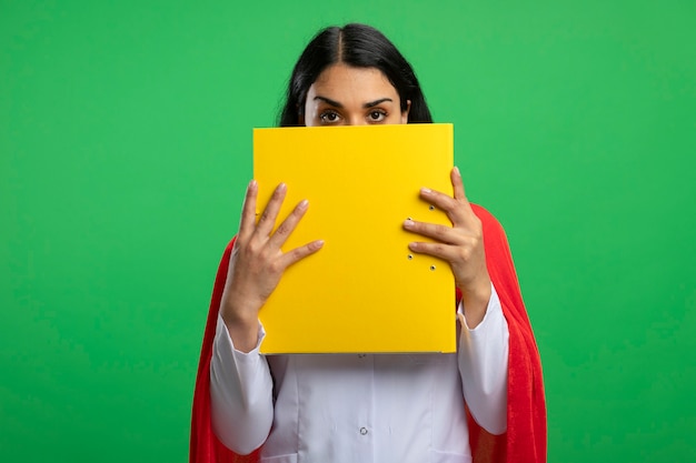 Free photo looking straight ahead young superhero girl wearing medical robe with stethoscope covered face with folder isolated on green