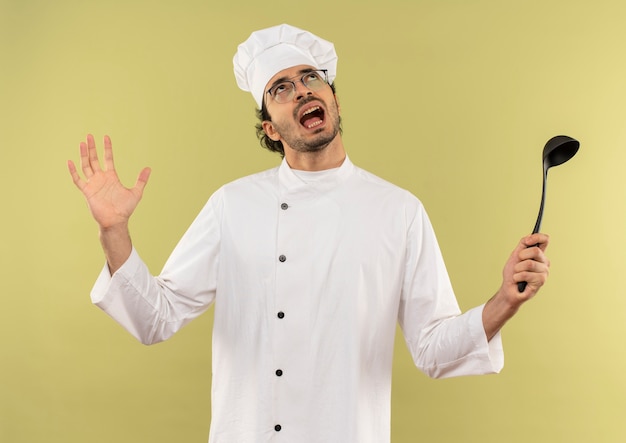 Foto gratuita guardando su arrabbiato giovane cuoco maschio che indossa l'uniforme del cuoco unico e bicchieri tenendo il mestolo e diffondere la mano