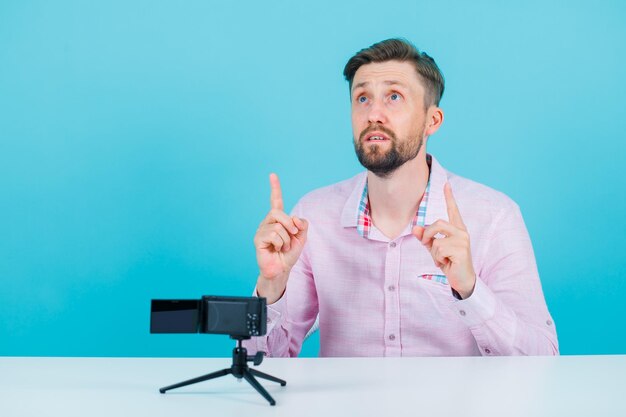 Looking up blogger ma is pointing up with forefingers by sitting in front of his mini camera on blue background