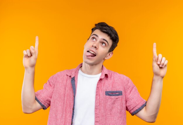 Looking to up caucasian young guy wearing pink shirt points to up showing tongue on isolated orange background