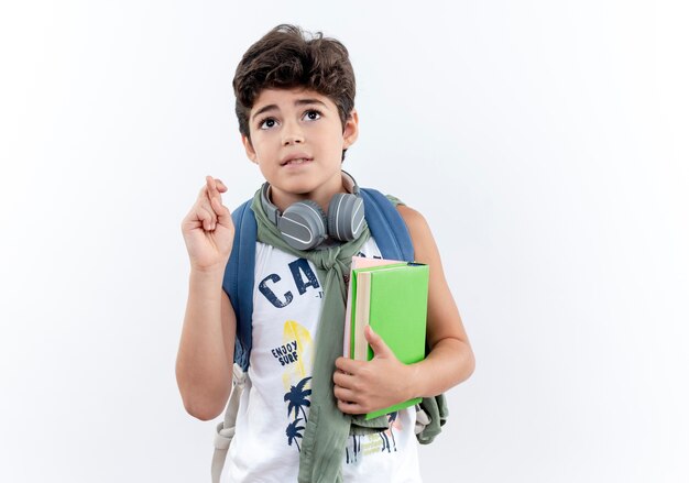 Looking at up concerned little schoolboy wearing back bag and headphones holding book and crossing finger isolated on white background