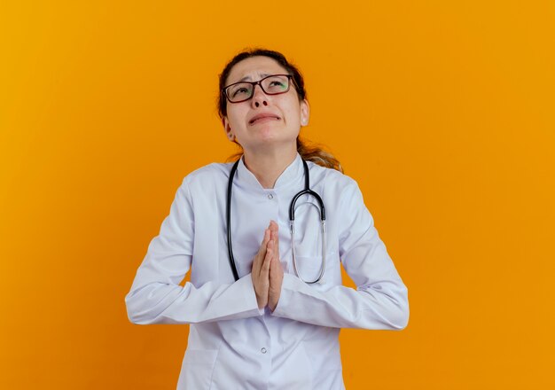 分離された祈りのジェスチャーを示す眼鏡と医療ローブと聴診器を身に着けている心配している若い女性医師を見上げる