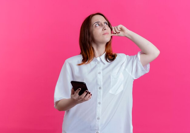 Looking at up confused young redhead girl holding phone and putting finger on head isolated on pink background