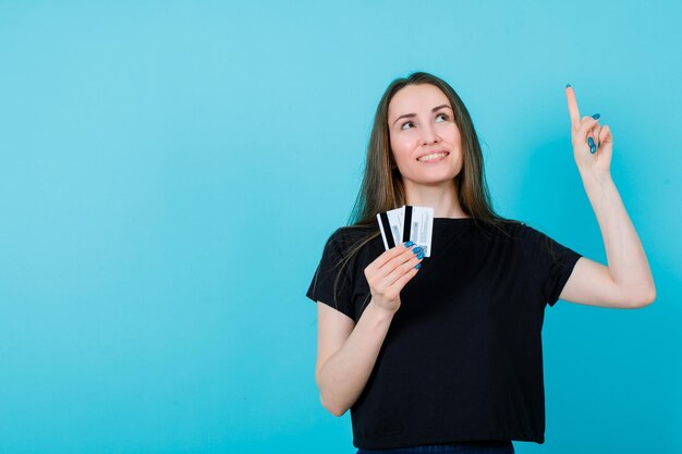 Looking up girl is pointing up with forefinger and holding credit cards on blue background
