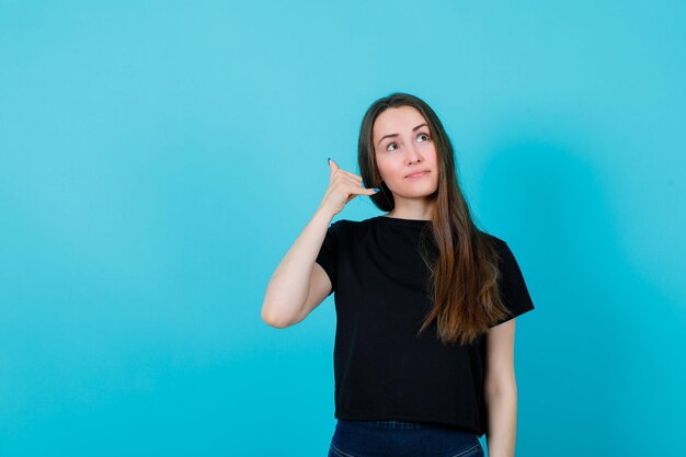 Looking up girl is showing phone gesture by holding hand near ear on blue background