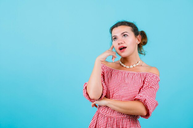 Looking up girl is thinking by holding forefinger on temple on blue background