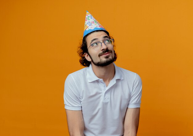 Looking at up impressed handsome man wearing glasses and birthday cap isolated on orange background