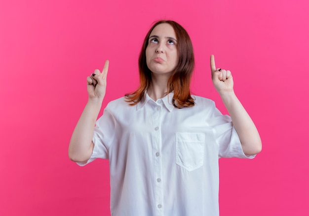 Looking at up impressed young redhead girl points at up isolated on pink wall