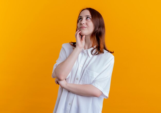 Looking at up impressed young redhead girl putting hand on cheek isolated on yellow background