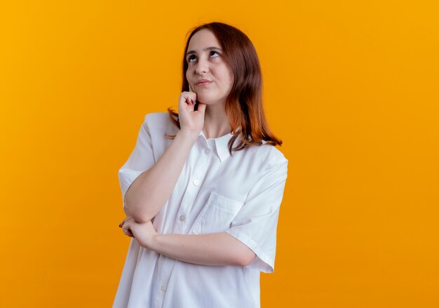 Looking at up impressed young redhead girl putting hand on cheek isolated on yellow background