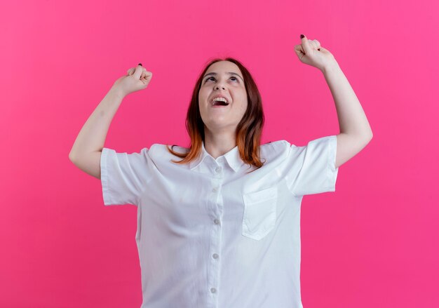 Looking at up joyful young redhead girl raising hands isolated on pink background