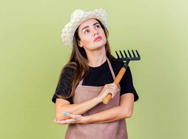 Looking at up pleased beautiful gardener girl in uniform wearing gardening hat holding rake isolated on olive green background