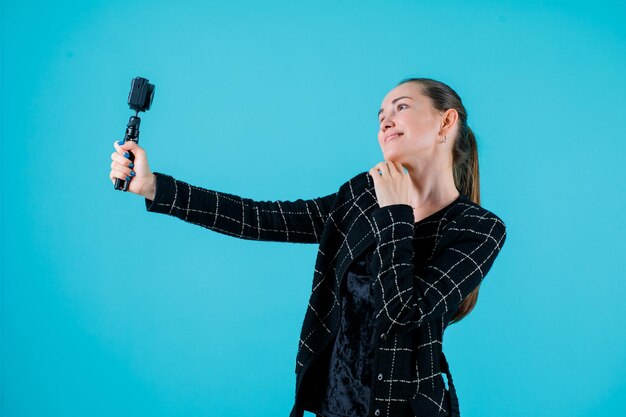 Looking up putting hand under chin and taking selfie with mini camera on blue background