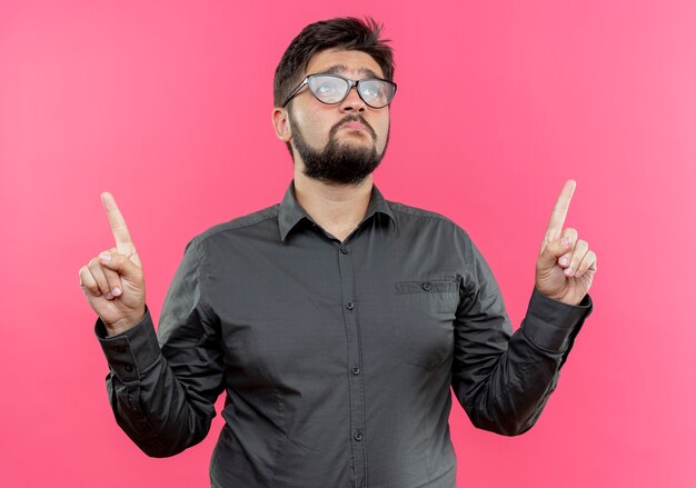 Looking at up sad young businessman wearing glasses points at up isolated on pink wall with copy space