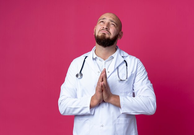 ピンクの壁に分離された祈りのジェスチャーを示す医療ローブと聴診器を身に着けている悲しい若い男性医師を見上げる