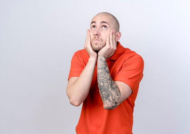 Looking at up sad young sporty man putting hands under chin isolated on white background with copy space