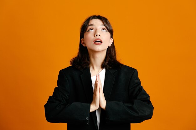 Looking at up showing pray gesture young beautiful female wearing black jacket isolated on orange background