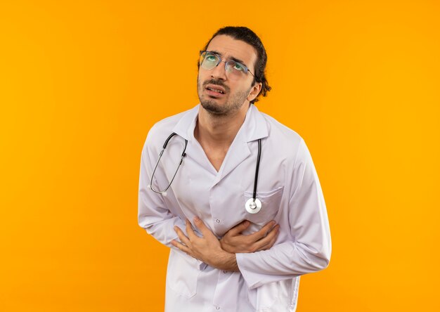 Looking at up sick young doctor with medical glasses wearing medical robe with stethoscope grabbed stomach