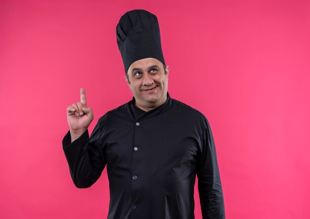 Looking at up smiling middle-aged male cook in chef uniform points finger to up on isolated pink wall with copy space