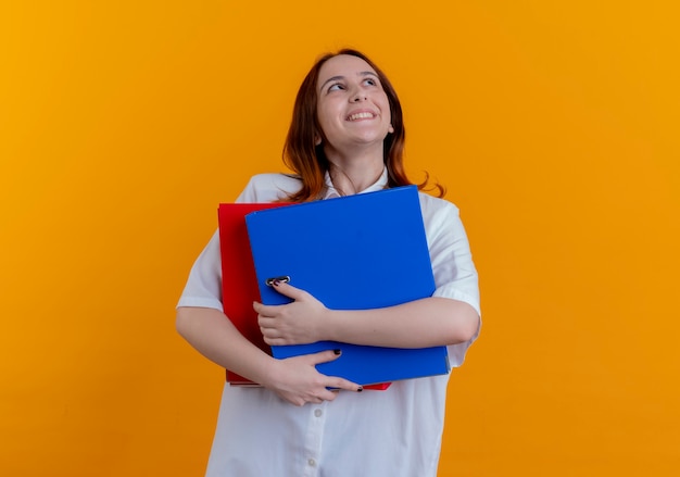 Looking at up smiling young redhead girl holding folders isolated on yellow background