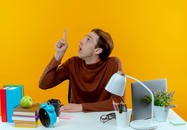 looking at up surprised young student boy sitting at desk with school tools points at up