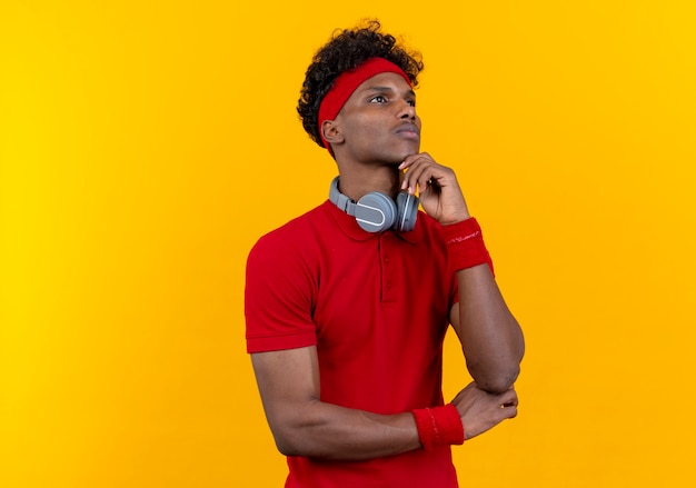 Looking at up thinking young afro-american sporty man wearing headband and wristband with headphones on shoulder putting hand under chin isolated on yellow background