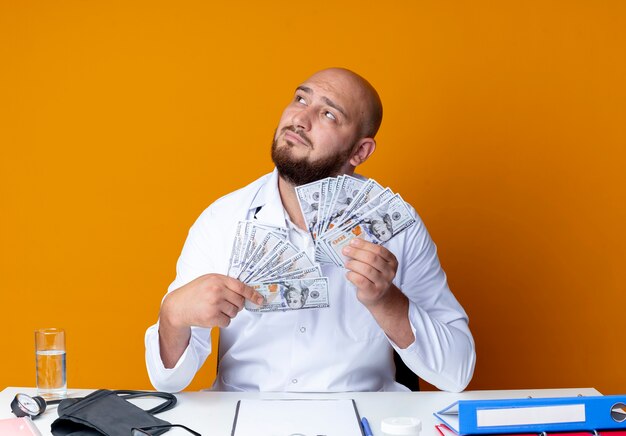 Foto gratuita guardando il pensiero di un giovane medico maschio calvo che indossa una tunica medica e uno stetoscopio seduto alla scrivania del lavoro