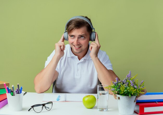 Looking at up thinking young handsome male student sitting at desk with school tools listen on headphones isolated on olive green