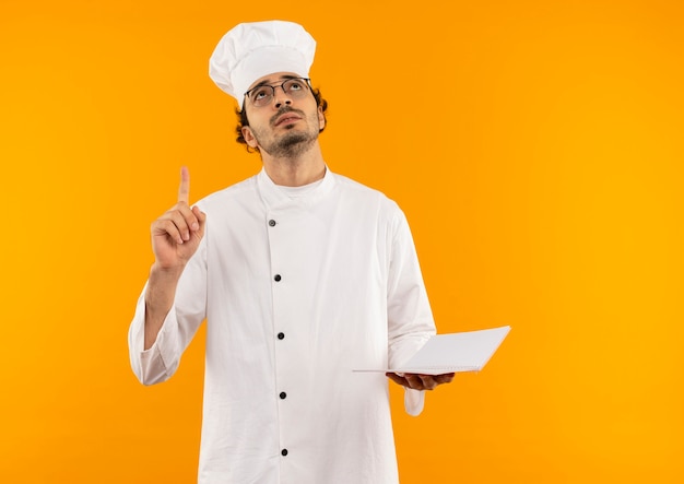 Looking at up thinking young male cook wearing chef uniform and glasses holding notebook and points to up 