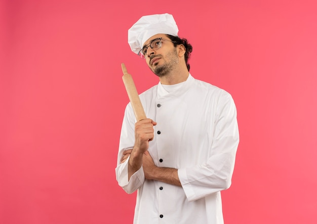 Looking at up thinking young male cook wearing chef uniform and glasses holding rolling pan 