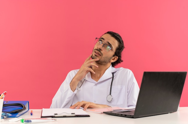 Looking at up thinking young male doctor with medical glasses wearing medical robe with stethoscope sitting at desk