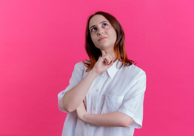 Looking at up thinking young redhead girl putting finger on neck isolated on pink background