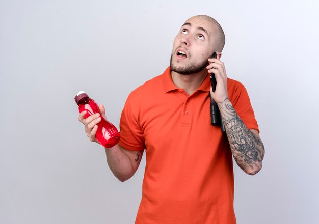 Looking at up thinking young sporty man holding water bottle and speaks on phone 