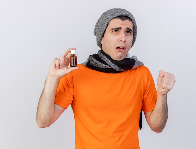 Foto gratuita guardando il debole giovane uomo malato che indossa il cappello invernale con sciarpa tenendo la medicina in bottiglia di vetro